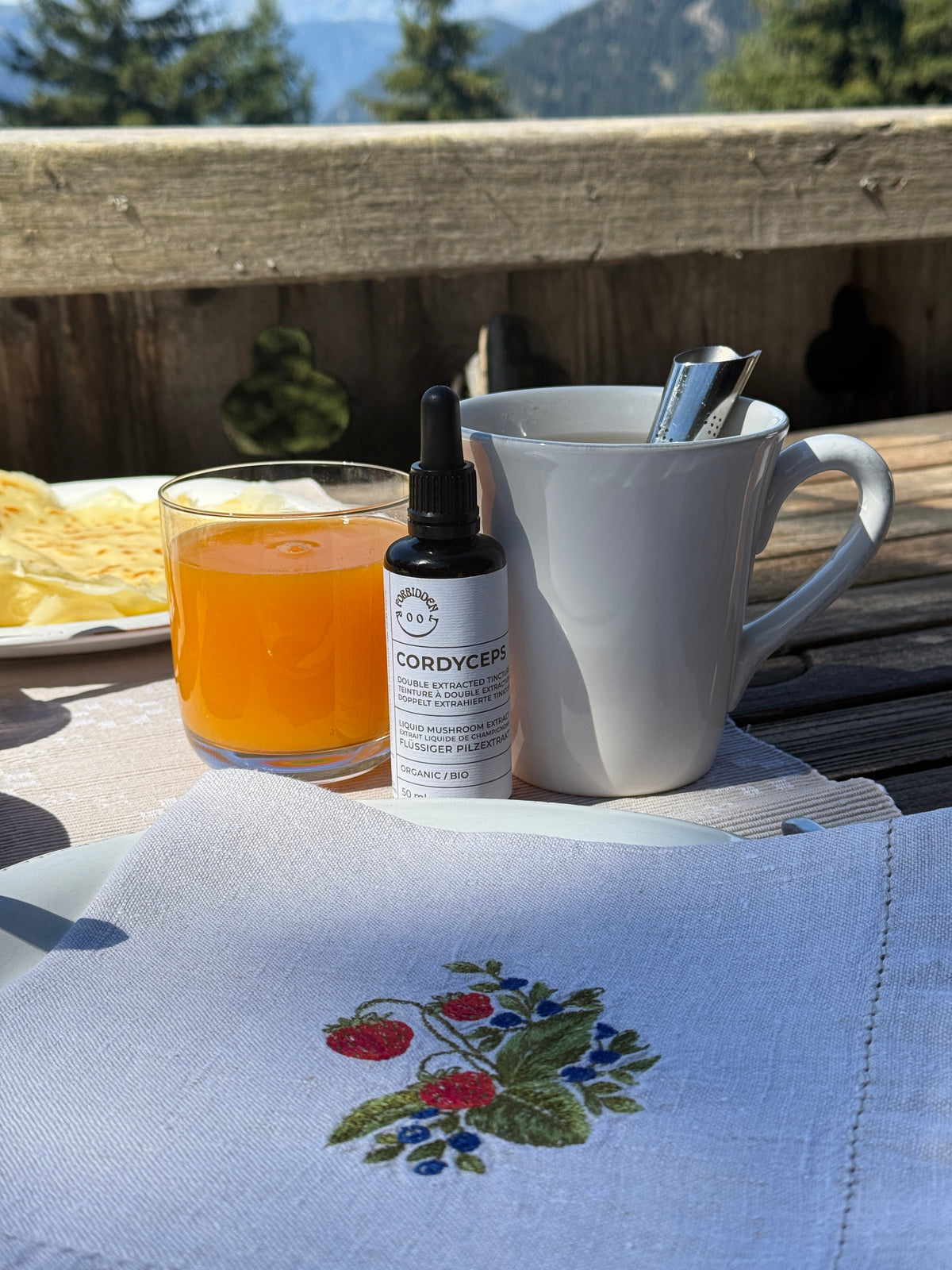 A small bottle of Cordyceps liquid supplement with a dropper cap sits on a wooden table outdoors, styled with a white mug, a glass of orange juice, and a beige embroidered napkin in the foreground.