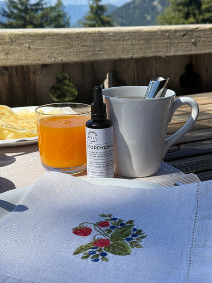 A small bottle of Cordyceps liquid supplement with a dropper cap sits on a wooden table outdoors, styled with a white mug, a glass of orange juice, and a beige embroidered napkin in the foreground.