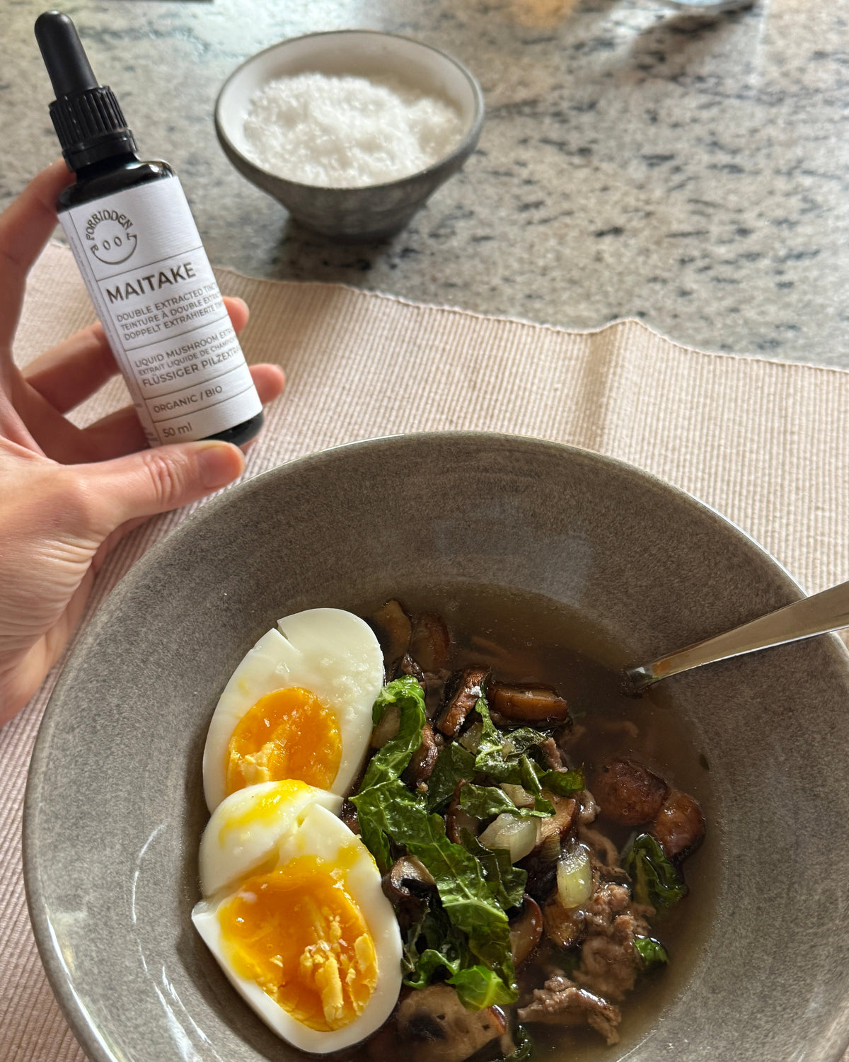 A hand holds a small black dropper bottle labeled MAITAKE over a bowl of soup topped with two halves of a soft-boiled egg. A small bowl of sea salt sits in the background on a speckled countertop.
