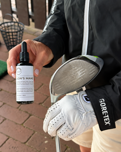 A person at a driving range holds a small bottle labeled 'LION'S MANE' in one hand while holding a golf club with a white GORE-TEX glove on the other hand. The person is wearing a black jacket and light-colored shorts.